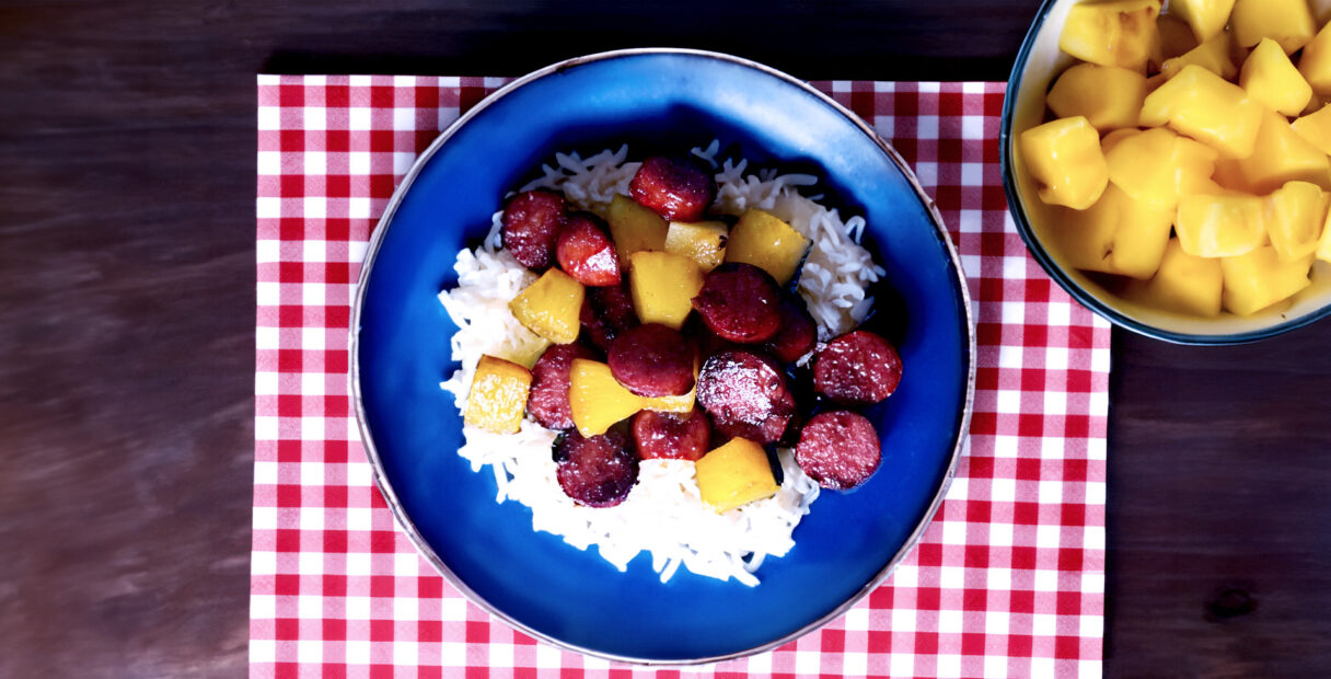 Delicious Hawaiian Kielbasa on a blue plate with rice and a red placemat and a bowl of pineapple