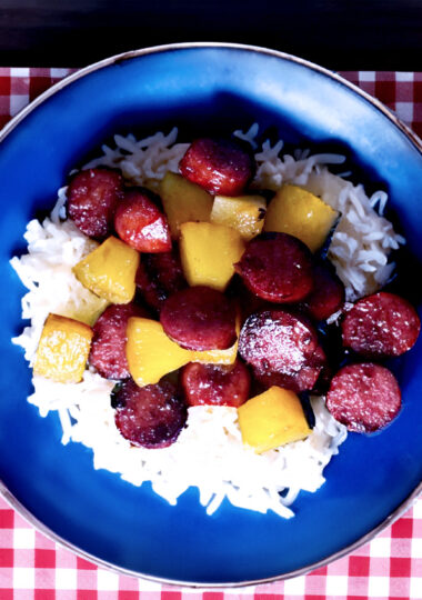 Delicious Hawaiian Kielbasa on a blue plate with rice and a red placemat and a bowl of pineapple