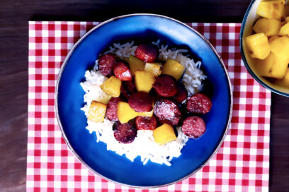 Delicious Hawaiian Kielbasa on a blue plate with rice and a red placemat and a bowl of pineapple