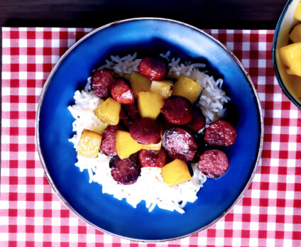 Delicious Hawaiian Kielbasa on a blue plate with rice and a red placemat and a bowl of pineapple